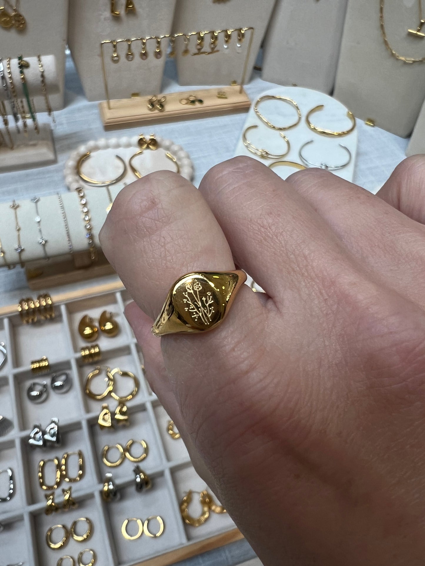 A gold signet ring with an engraved floral motif worn on a person's hand, set against a background of various jewelry pieces displayed on stands and trays.