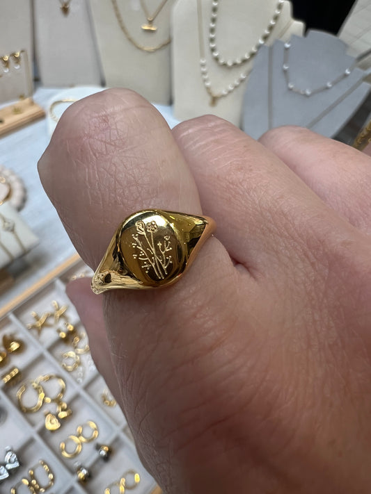 A gold signet ring with an engraved floral motif on a person's finger, with jewelry displays visible in the blurred background.