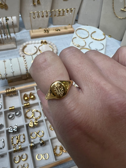 A gold signet ring with an engraved floral motif worn on a person's hand, set against a background of various jewelry pieces displayed on stands and trays.